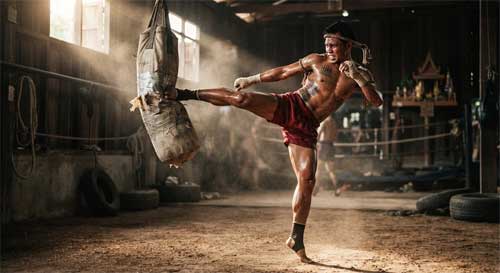 Kaensak ejecutando una patada circular alta con precisión milimétrica contra un saco pesado, mostrando la tensión muscular y la técnica perfecta del Muay Thai tradicional